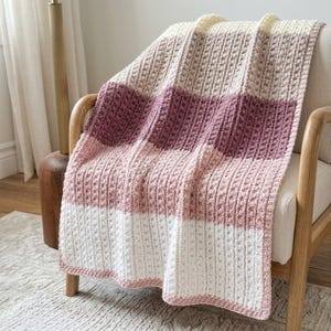 Può includere: Una coperta all'uncinetto con strisce orizzontali a blocchi di colore crema, malva e rosa chiaro. Il tessuto presenta un motivo costante a punto nocciolina in rilievo, creando una superficie strutturata sull'intero pezzo rettangolare.