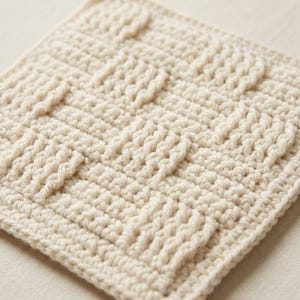 Op de afbeelding: Een vierkant gehaakt stuk in een off-white kleur met een getextureerd mandvlechtpatroon. Het ontwerp bestaat uit verhoogde verticale en horizontale steken die een rasterachtig uiterlijk creëren over het gehele oppervlak van de stof.