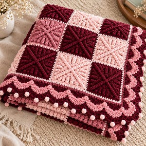 May include: A crocheted blanket featuring a checkerboard pattern of deep burgundy and light pink squares. Each square has a raised cross design. The blanket is finished with a scalloped border in burgundy and pink with small pom-poms.