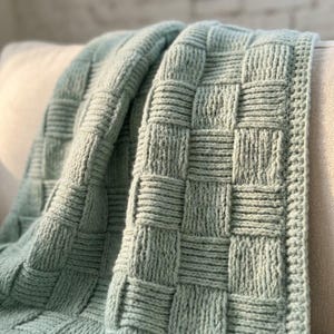 May include: A sage green knitted throw blanket featuring a prominent basketweave texture. The design consists of alternating vertical and horizontal rectangular blocks of knit stitches, creating a raised, grid-like pattern across the entire surface.