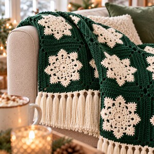 May include: A dark forest green crochet throw blanket featuring a repeating pattern of cream-colored snowflake motifs in each square. The blanket is finished with a long, cream-colored fringe along the bottom edge, adding texture to the design.