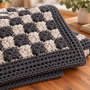 May include: A crocheted blanket featuring a checkerboard pattern of dark charcoal gray and cream-colored squares. The blanket is finished with a wide, solid dark charcoal gray crocheted border that frames the entire square-patterned center.