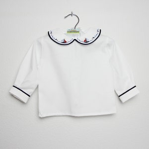 May include: White long-sleeved shirt with a Peter Pan collar. The collar is decorated with embroidered sailboats in red, blue, and white. The cuffs and collar are trimmed with navy blue piping. The shirt is hanging on a silver hanger.