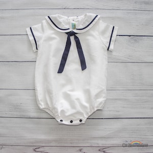 May include: White baby romper with a navy blue sailor collar and tie. The short-sleeved romper has a rounded neckline and snaps at the bottom. The outfit is made of a soft fabric and is designed for infants.