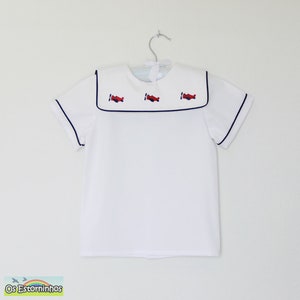 Puede incluir: Camisa blanca de manga corta con cuello marinero. El cuello está ribeteado con ribete azul marino y presenta tres aviones rojos bordados. La camisa está colgada de una percha de metal.