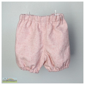 May include: Pink baby shorts with an elastic waistband and gathered leg openings. The shorts are made of a textured fabric with a subtle flecked pattern. The garment is hanging from a clothesline against a white background.