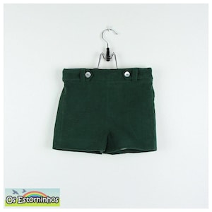 Könnte beinhalten: Dunkelgrüne Shorts mit zwei weißen Knöpfen am Bund, hängend an einem Metallbügel. Die Shorts sind aus einem strukturierten Stoff gefertigt. Unten links im Bild befindet sich das Logo "Os Estorninhos".