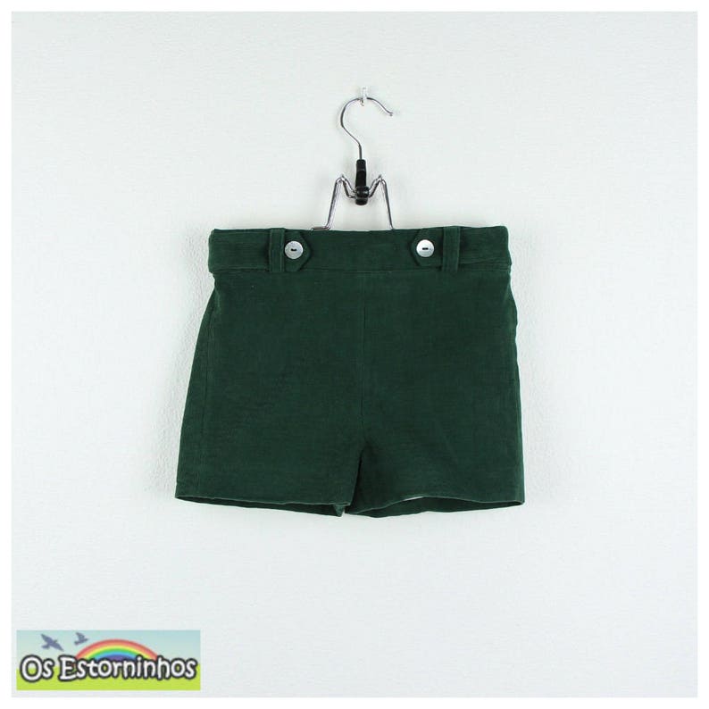May include: Dark green shorts with two white buttons on the waistband, hanging from a metal hanger. The shorts are made of a textured fabric. The bottom left corner of the image has a small logo with the text "Os Estorninhos".