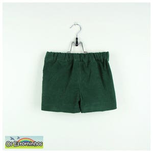 May include: Dark green corduroy shorts hanging on a metal hanger. The shorts have an elastic waistband. The label at the bottom left corner reads "Os Estorninhos" with a small bird graphic.
