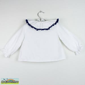 Op de afbeelding: Witte blouse met lange mouwen en een gerimpelde kraag afgezet met marineblauwe pompons. De blouse heeft een eenvoudig ontwerp met gerimpelde manchetten en hangt aan een metalen hanger. De blouse is waarschijnlijk voor een kind.