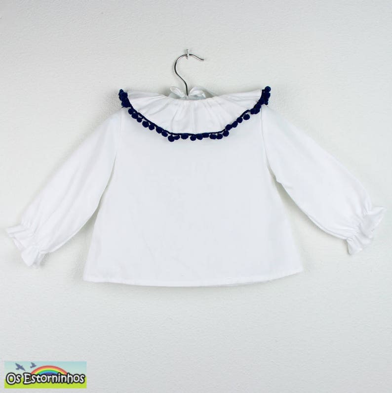 Op de afbeelding: Witte blouse met lange mouwen en een gerimpelde kraag afgezet met marineblauwe pompons. De blouse heeft een eenvoudig ontwerp met gerimpelde manchetten en hangt aan een metalen hanger. De blouse is waarschijnlijk voor een kind.