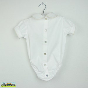 May include: White baby bodysuit with a Peter Pan collar and short sleeves. The collar and sleeve edges have a light beige trim. The bodysuit has a button-down front with six pearlescent buttons and is hanging on a metal hanger.