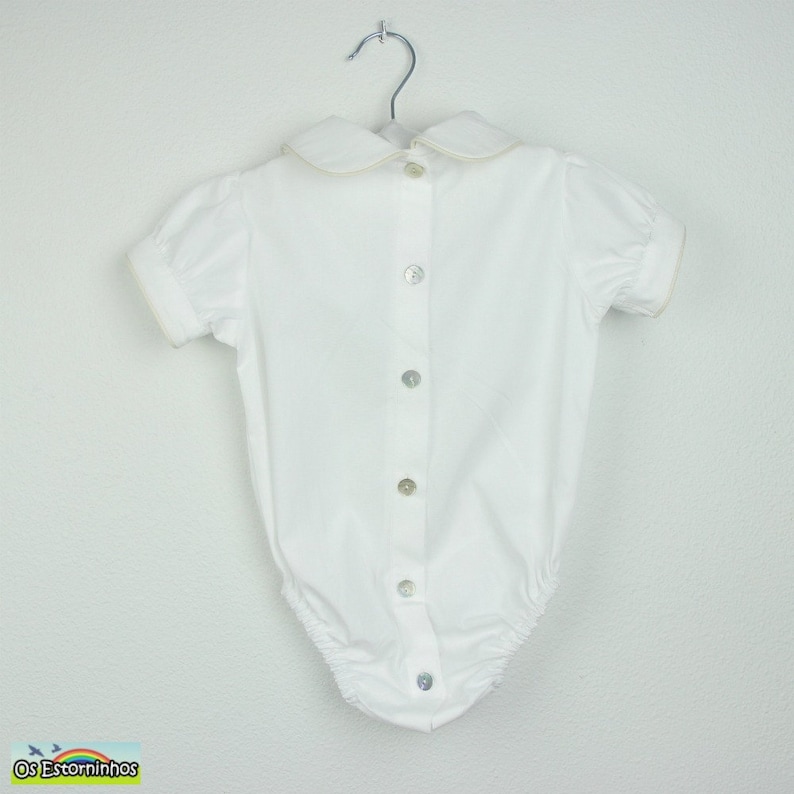 May include: White baby bodysuit with a Peter Pan collar and short sleeves. The collar and sleeve edges have a light beige trim. The bodysuit has a button-down front with six pearlescent buttons and is hanging on a metal hanger.
