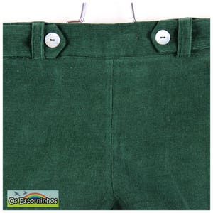 May include: Close-up of dark green corduroy pants. The pants have a high waist with two white buttons and a button closure. The fabric has a ribbed texture. The brand name "Os Estorninhos" is visible in a small label at the bottom.