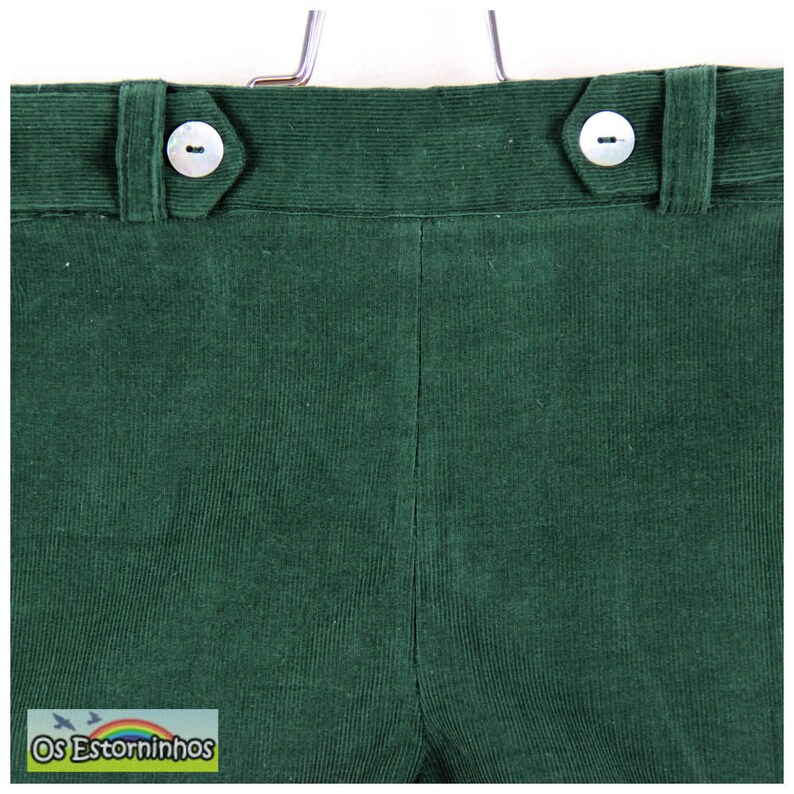 May include: Close-up of dark green corduroy pants. The pants have a high waist with two white buttons and a button closure. The fabric has a ribbed texture. The brand name "Os Estorninhos" is visible in a small label at the bottom.