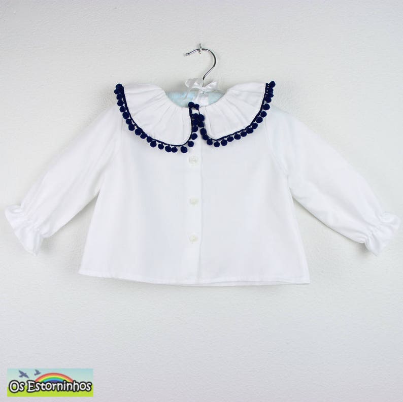 Op de afbeelding: Witte blouse met lange mouwen en een kraag met marineblauwe pompons. De blouse heeft een knoopsluiting aan de voorkant en gerimpelde manchetten. Opgehangen aan een zilveren hanger.