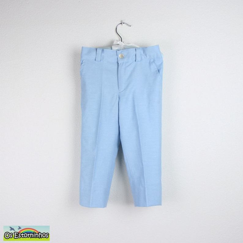 May include: Light blue pants hanging on a white background. The pants have a button closure, belt loops, and pockets. The fabric has a subtle texture. The pants are likely for children.