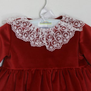 May include: A red velvet dress with a white lace collar. The collar is adorned with a delicate floral pattern. The dress is hanging on a hanger with the text "Os Estorninhos".