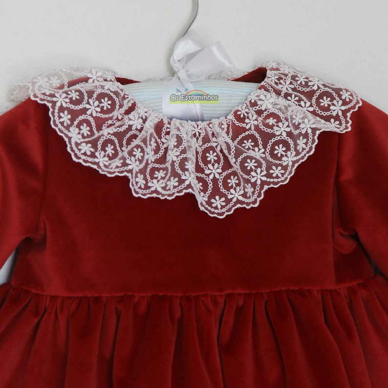 May include: A red velvet dress with a white lace collar. The collar is adorned with a delicate floral pattern. The dress is hanging on a hanger with the text "Os Estorninhos".