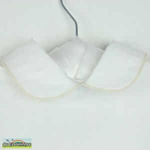 May include: A white Peter Pan collar with a cream-colored trim, hanging from a gray hanger. The collar is made of a smooth, crisp fabric. The bottom left corner of the image has the text "Os Estorninhos" with a rainbow and birds.