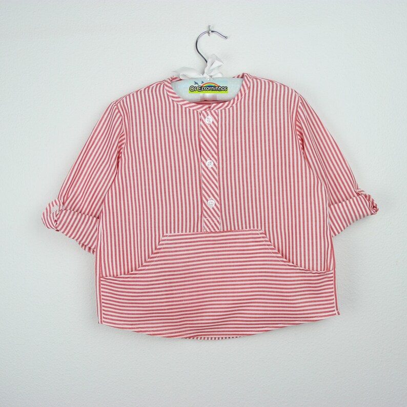 May include: A red and white striped shirt with a mandarin collar and a front pocket. The long-sleeved shirt has rolled-up cuffs and a button-down placket. The shirt is hanging on a metal hanger.