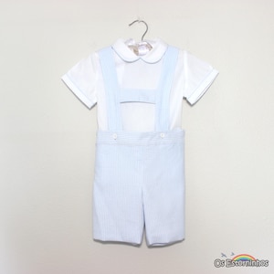 May include: A white short-sleeved shirt and light blue striped shorts set for a child. The shirt has a rounded collar and light blue accents. The shorts have suspenders attached. The outfit is hanging on a hanger.