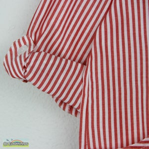 May include: Close-up of a red and white striped fabric, likely a shirt or dress. The sleeve is rolled up and buttoned, showcasing the interior white fabric. The fabric has vertical stripes. The brand name "Os Estorninhos" is visible.