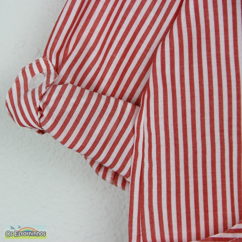 May include: Close-up of a red and white striped fabric, likely a shirt or dress. The sleeve is rolled up and buttoned, showcasing the interior white fabric. The fabric has vertical stripes. The brand name "Os Estorninhos" is visible.