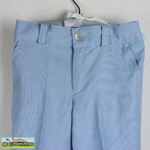 May include: Light blue dress pants with a button closure and belt loops. The pants have two front pockets and are hanging on a hanger. The fabric has a subtle texture. The brand name "Os Estorninhos" is visible in the bottom left corner.