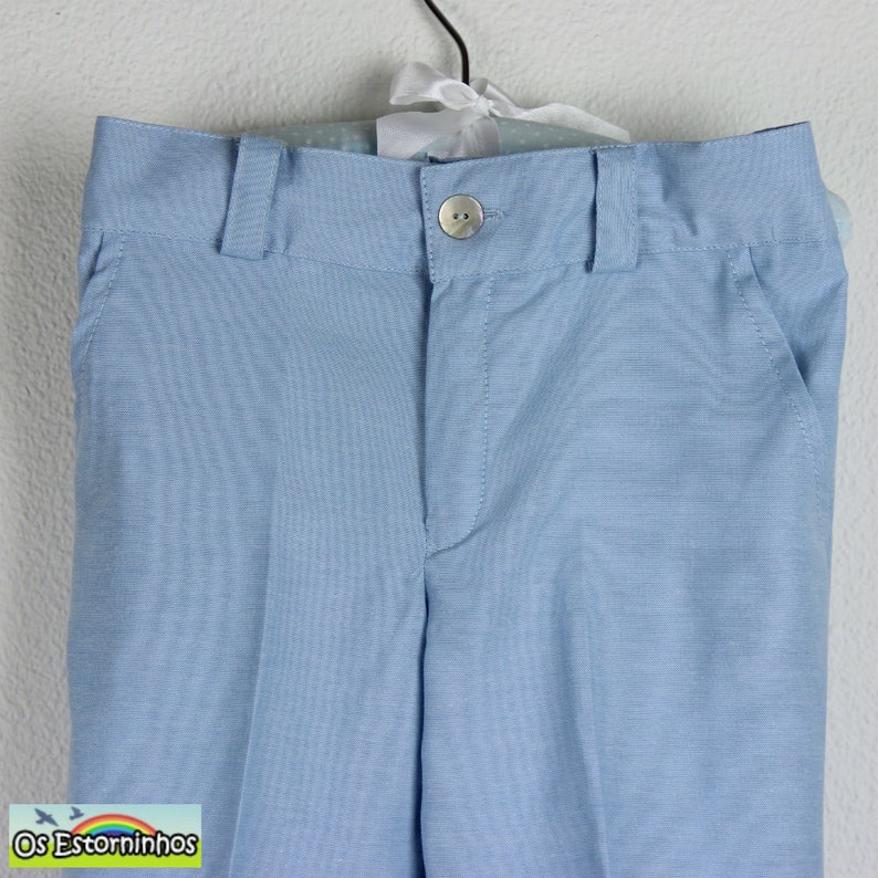 May include: Light blue dress pants with a button closure and belt loops. The pants have two front pockets and are hanging on a hanger. The fabric has a subtle texture. The brand name "Os Estorninhos" is visible in the bottom left corner.