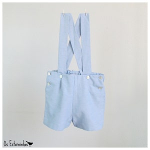 May include: Light blue baby shorts with suspenders, featuring white buttons and a comfortable elastic waistband. The suspenders are attached to the shorts with white buttons. The shorts are hanging on a clothesline.