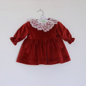 May include: A red velvet dress with a white lace collar and long sleeves. The dress is hanging on a silver hanger against a white background. The sleeves have ruffled cuffs.