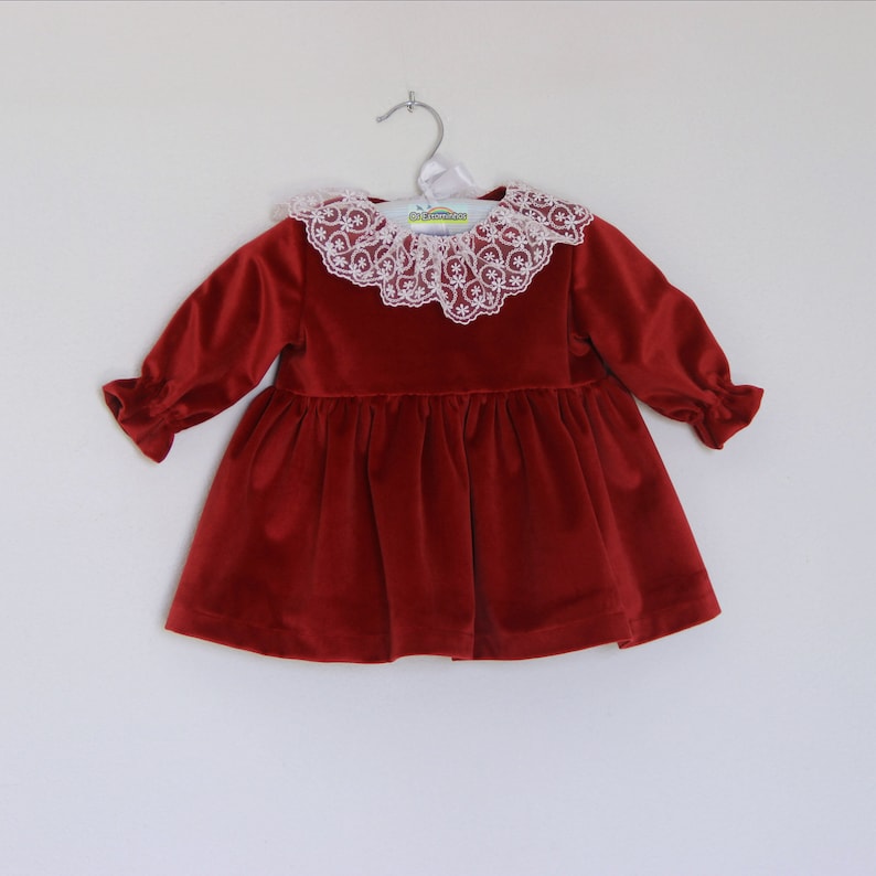 May include: A red velvet dress with a white lace collar and long sleeves. The dress is hanging on a silver hanger against a white background. The sleeves have ruffled cuffs.
