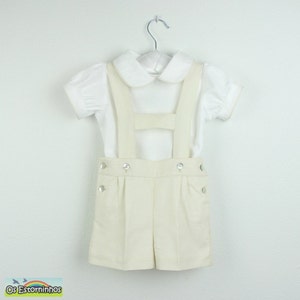 May include: A cream-colored shorts set with suspenders and a white short-sleeved shirt. The shorts have decorative buttons. The outfit is hanging on a metal hanger. The set is designed for children.