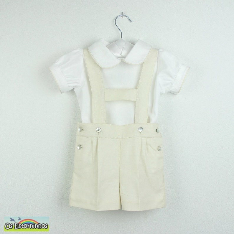 May include: A cream-colored shorts set with suspenders and a white short-sleeved shirt. The shorts have decorative buttons. The outfit is hanging on a metal hanger. The set is designed for children.