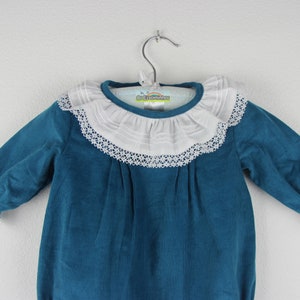 May include: A teal corduroy baby romper with long sleeves and a white lace-trimmed collar. The collar is a prominent feature, adding a touch of elegance to the garment. The romper is hanging on a metal hanger.