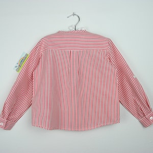 May include: A red and white striped long-sleeved shirt, hanging on a metal hanger. The shirt has a button-down front and buttoned cuffs. The back of the shirt is visible, showing vertical stripes and a tag with text.