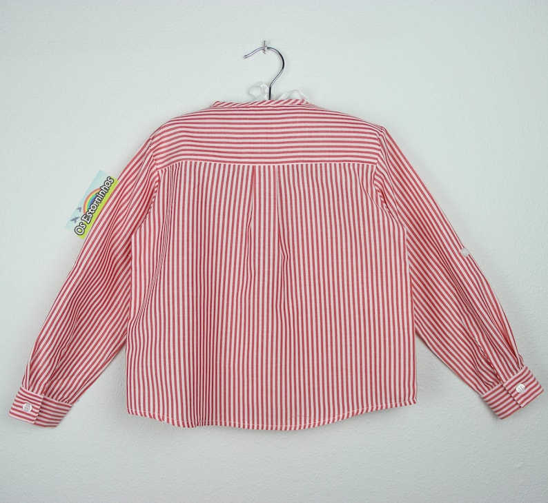May include: A red and white striped long-sleeved shirt, hanging on a metal hanger. The shirt has a button-down front and buttoned cuffs. The back of the shirt is visible, showing vertical stripes and a tag with text.
