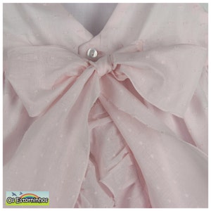 May include: A light pink baby dress with a large bow. The dress features a V-neckline and a button closure. The bow is made of a sheer fabric with a dotted pattern. The dress has a ruffled design.