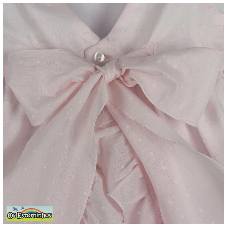 May include: A light pink baby dress with a large bow. The dress features a V-neckline and a button closure. The bow is made of a sheer fabric with a dotted pattern. The dress has a ruffled design.
