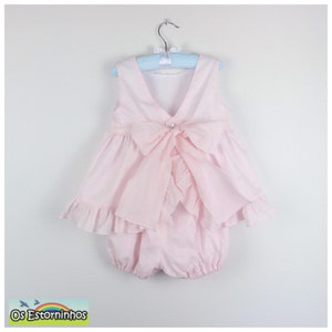 May include: A light pink baby romper with a large bow on the back and a pearl button closure. The romper has a ruffled skirt and is hanging on a blue hanger. The brand name "Os Estorninhos" is visible in the bottom left corner.