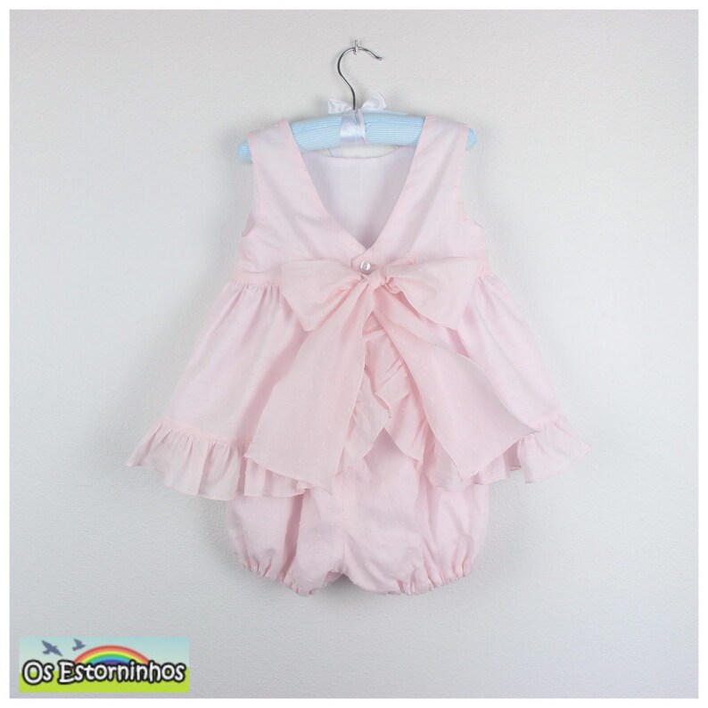 May include: A light pink baby romper with a large bow on the back and a pearl button closure. The romper has a ruffled skirt and is hanging on a blue hanger. The brand name "Os Estorninhos" is visible in the bottom left corner.