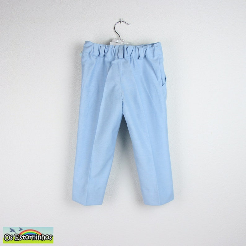 May include: Light blue pants hanging on a metal hanger. The pants have an elastic waistband and pockets. The fabric appears to be a lightweight cotton or linen blend. The pants are designed for children.