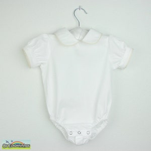 May include: White baby bodysuit with short, puffed sleeves and a Peter Pan collar. The collar and sleeve edges have a delicate gold trim. The bodysuit has snap closures at the bottom and is hanging on a silver hanger.