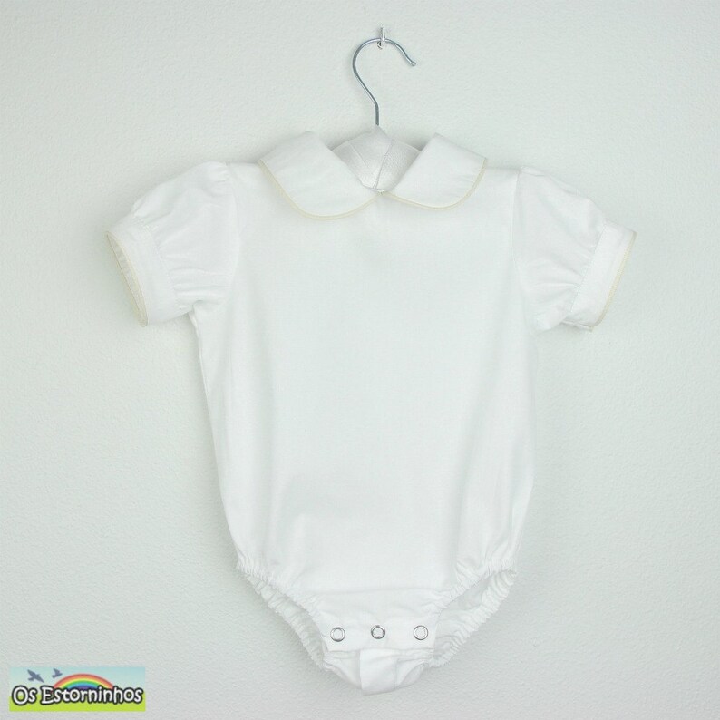May include: White baby bodysuit with short, puffed sleeves and a Peter Pan collar. The collar and sleeve edges have a delicate gold trim. The bodysuit has snap closures at the bottom and is hanging on a silver hanger.