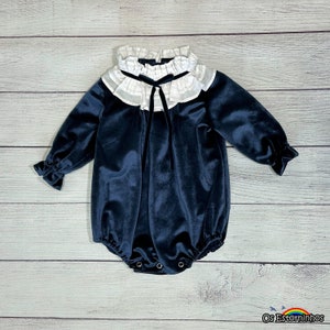 May include: A navy blue velvet baby romper with long sleeves and a white ruffled collar. The romper has a black bow at the neckline and snaps at the bottom. The sleeves have ruffled cuffs. The background is a light wood grain.