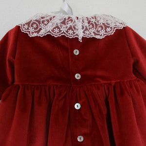 May include: A deep red velvet dress with a white lace collar featuring a floral pattern. The dress has long sleeves and a gathered waist. Pearl buttons run down the back of the dress.
