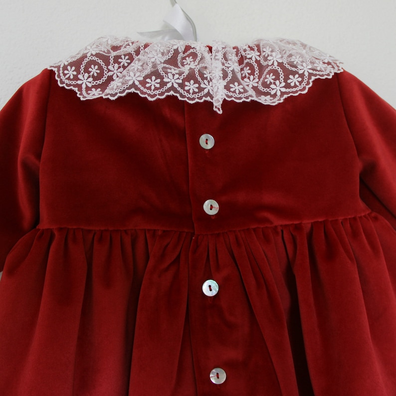 May include: A deep red velvet dress with a white lace collar featuring a floral pattern. The dress has long sleeves and a gathered waist. Pearl buttons run down the back of the dress.