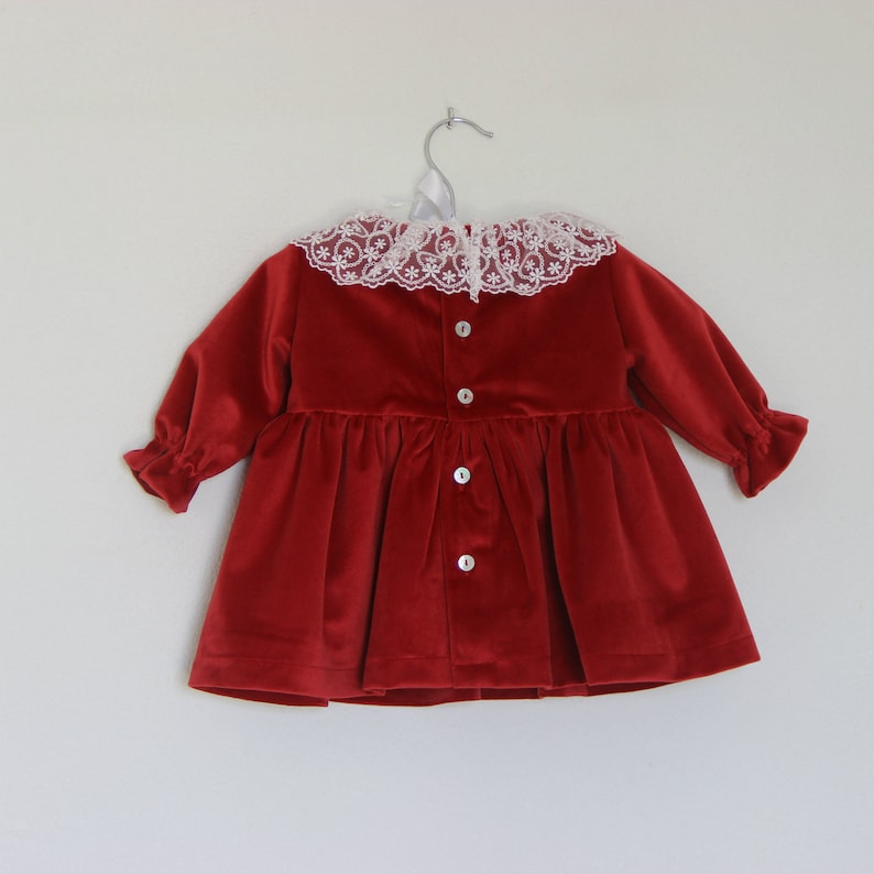 May include: A red velvet dress with a white lace collar and long sleeves. The dress features a button-down front and a gathered skirt. The dress is hanging on a silver hanger.