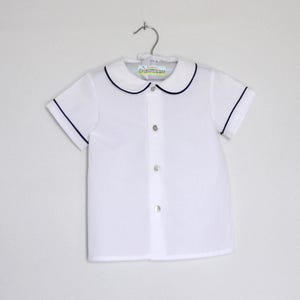 May include: White short-sleeved shirt with navy blue trim on the collar and sleeves. The shirt has a button-down front and is hanging on a metal hanger. The shirt is made of a lightweight fabric.